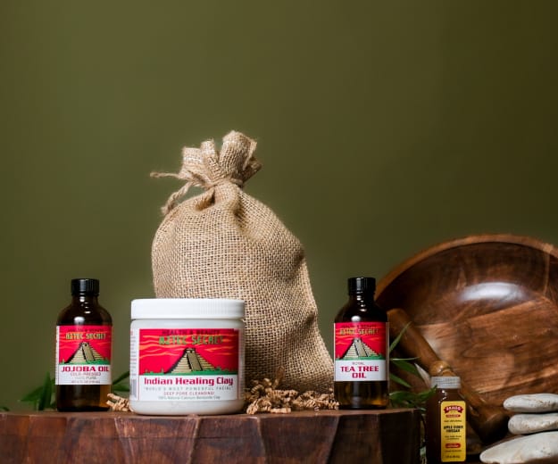 A collection of health and beauty products including Indian Healing Clay, Tea Tree Oil, and Jojoba Oil on a wooden surface with a green background.
