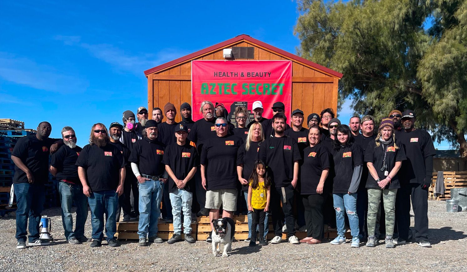 A group photo of a large number of people, both men and women, standing outside in front of a wooden building with a sign that says 'Health & Beauty Aztec Secret'. They are all wearing black shirts and appear to be a team or organization.